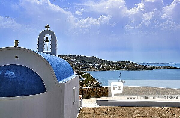 Traditionelle griechisch orthodoxe Kirche mit blauer Kuppel und Glockenturm mit Blick auf die typische griechische Küsten oder Insellandschaft an einem Sommertag. Einsame Segelboot navigieren die Bucht. Grüne Hügel  ruhige Hafengewässer  blauer Himmel mit einigen Wolken. Gelassenheit  Urlaub