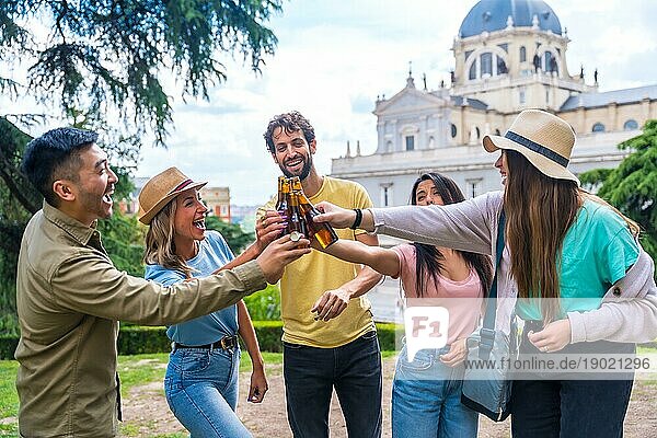 Eine multiethnische Gruppe von Freunden feiert in einem Stadtpark mit Bier. Lächelnd und mit den Flaschen anstoßend