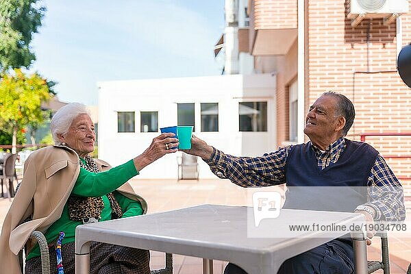 Zwei ältere Menschen im Garten eines Pflegeheims  die an einem sonnigen Sommertag mit leuchtenden Gläsern anstoßen