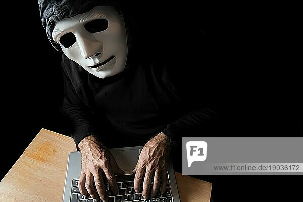 Schwarz gekleideter Hacker mit Maske und Kapuze tippt auf einem Computer  während er in die Kamera schaut CybercrimeKonzept