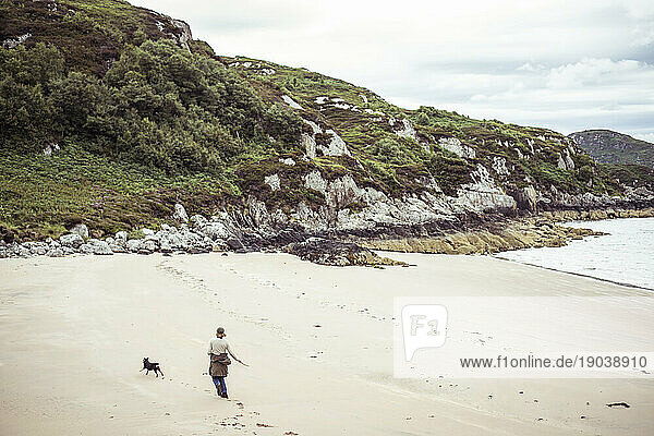 woman and dog run along remote empty beach on rugged coast of Scotland