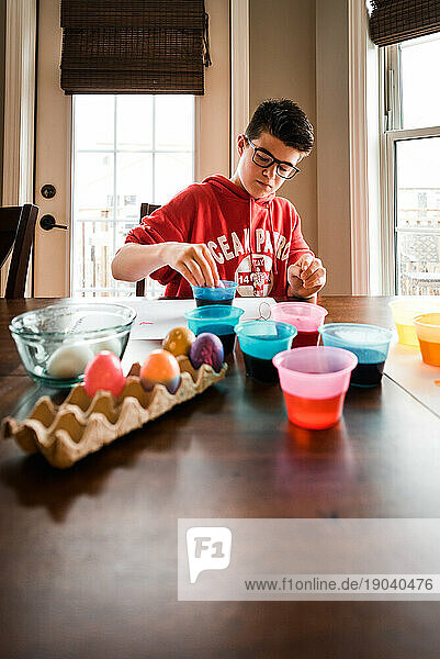 Teenage boy dying Easter eggs with containers of dye at the table.