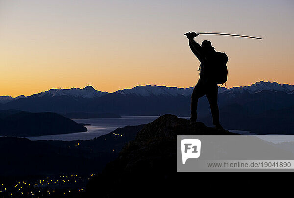 A Man With A Bamboo Stick Swings It Above His Head While Standing On A Mountain Peak During Sunset