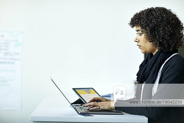 Side view of serious businesswoman using laptop computer on desk while standing against wall in office