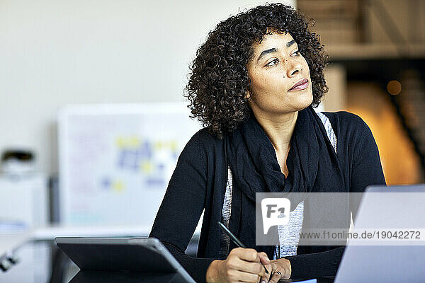 Thoughtful businesswoman with curly hair looking away while standing at desk in office