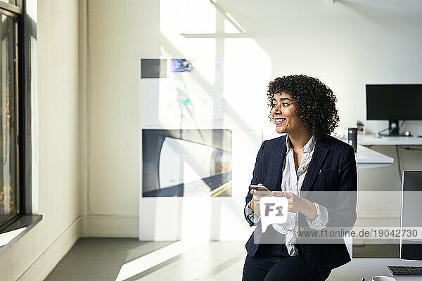 Smiling businesswoman with smart phone looking away while standing against wall in office