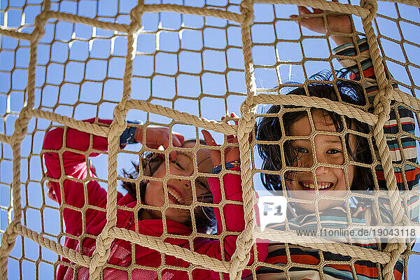 two laughing children peer down through a rope net against blue sky