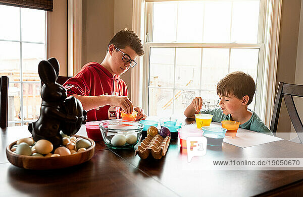 Two boys coloring Easter eggs with containers of dye at wooden table.