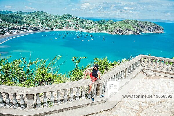 Glücklicher Tourist  der an einem Aussichtspunkt einer Bucht sitzt. Porträt des Touristen Mann in der Ansicht der Bucht von San Juan del Sur. Reisen und Tourismus Förderung Konzept