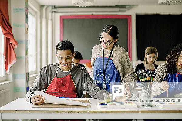 Lächelnder männlicher Jugendlicher  der einer Lehrerin während des Malkurses sein Kunstwerk zeigt