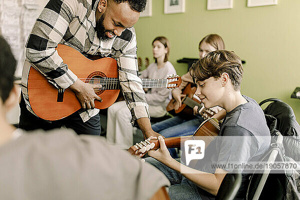 Junger männlicher Lehrer  der einem Teenager beim Gitarrenspiel im Musikunterricht an einer High School hilft