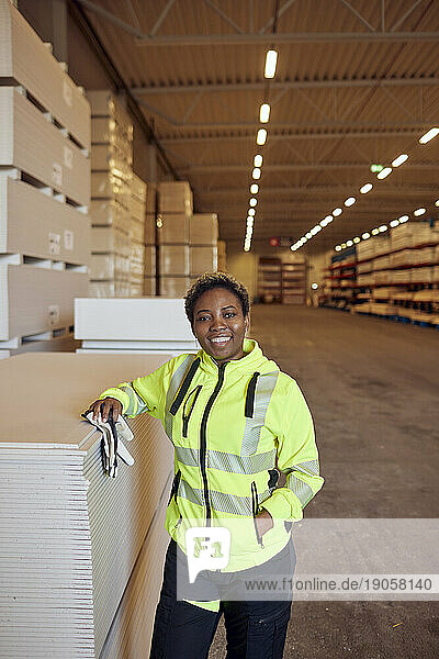 Porträt eines lächelnden Arbeiters in Arbeitsschutzkleidung  der sich in einer Fabrik auf Bretter stützt
