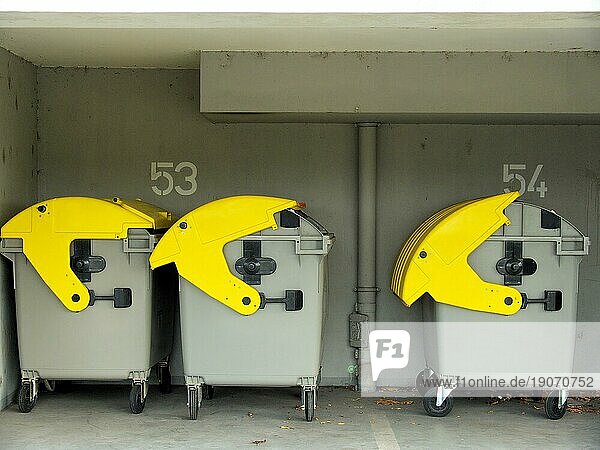 Drei fahrbare Restmüll-Container mit gelben Deckeln  unterschiedlich weit geöffnet  im Hintergrund die Zahlen 53 und 54 für die Wohneinheiten und ein Abflussrohr in grau