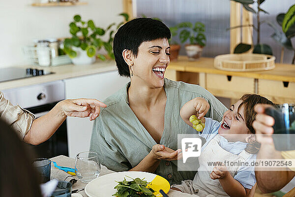 Smiling son holding grapes sitting on mother's lap at dining table in kitchen