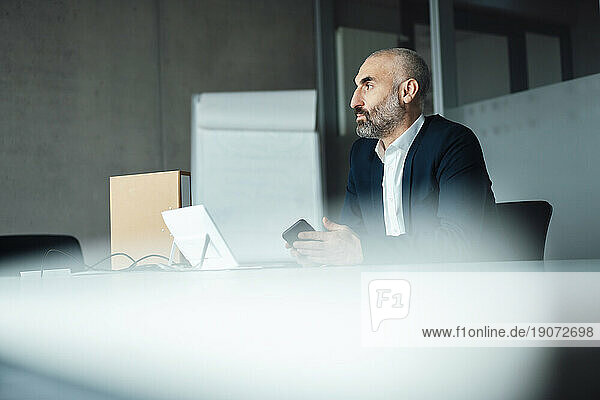 Thoughtful mature businessman sitting with smart phone at desk