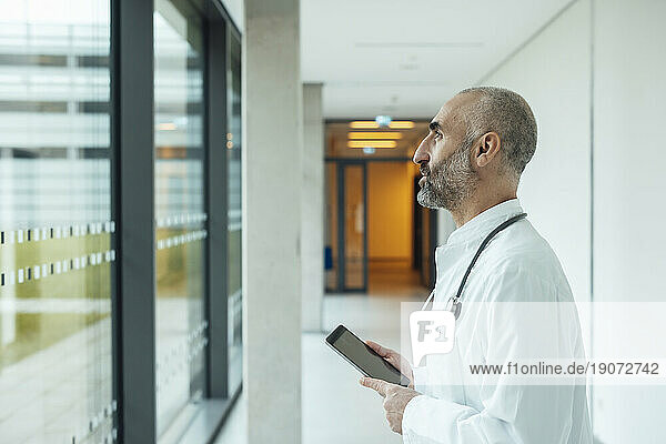 Thoughtful mature doctor with tablet PC looking through window in hospital