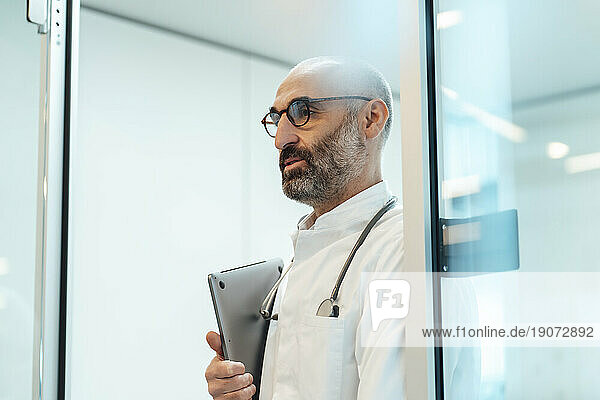 Thoughtful doctor standing with laptop in doorway