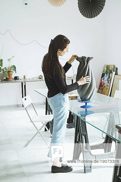 Young craftswoman looking at vase standing in workshop