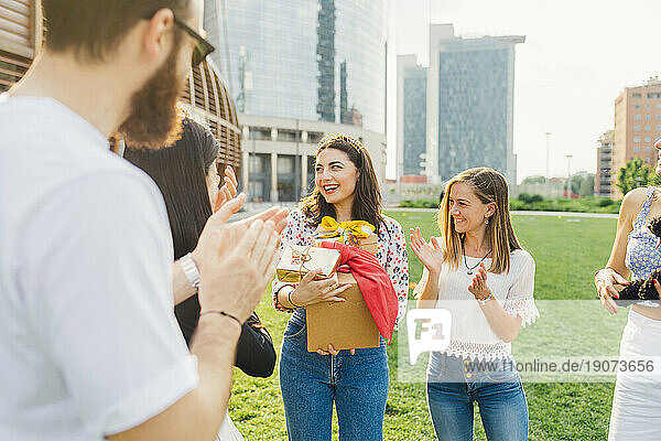 Happy woman holding gifts with friends applauding in park