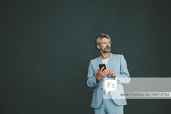 Thoughtful businessman with smart phone standing against teal background