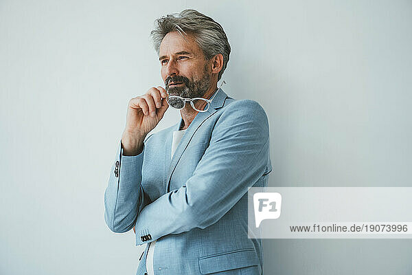 Thoughtful businessman holding eyeglasses in front of wall