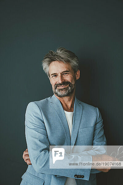Smiling businessman with arms crossed leaning on teal background