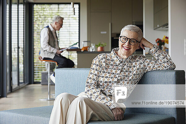 Portrait of smiling senior woman sitting on couch at home with man in background