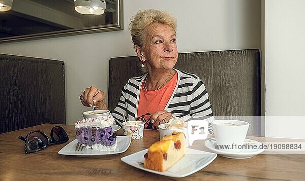 Attraktive  fröhliche ältere Frau trinkt Kaffee  während sie in einem Café sitzt. Porträt einer reifen Frau in einer Cafeteria. Bunte Torten und Kaffee auf dem Tisch. Angenehme entspannende Atmosphäre. Hübsche Frau mittleren Alters mit einer Tasse Kaffee