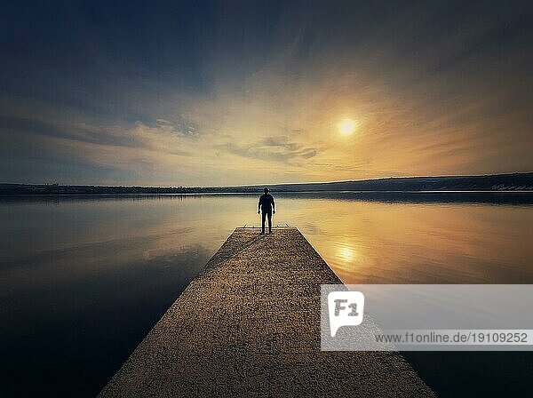 Mann  der allein auf dem Pier steht und den Sonnenuntergang betrachtet  der sich im ruhigen Wasser des Sees spiegelt. Scenic Ansicht mit einer einsamen Person auf dem Dock Blick auf den Horizont