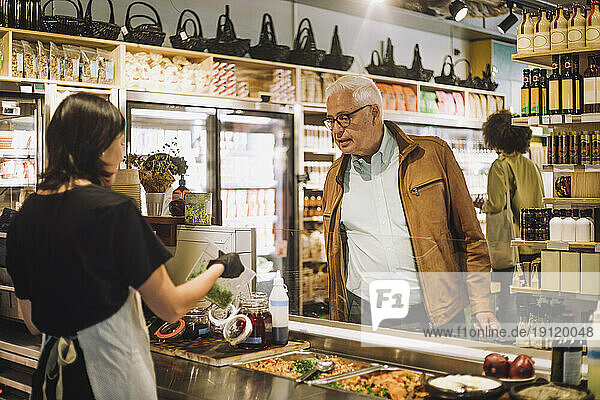 Verkäuferin zeigt einem älteren männlichen Kunden im Feinkostladen einen Behälter
