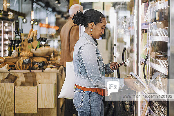 Side view of female customer photographing package while standing at grocery store