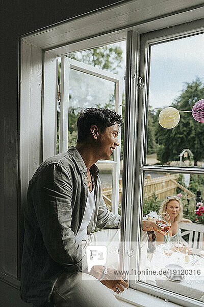 Lächelnder junger Mann  der auf der Fensterbank sitzt und seinen Freunden beim Feiern im Café zusieht