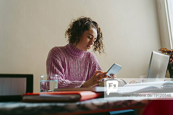 Junge Frau benutzt ihr Smartphone  während sie am Tisch Hausaufgaben macht