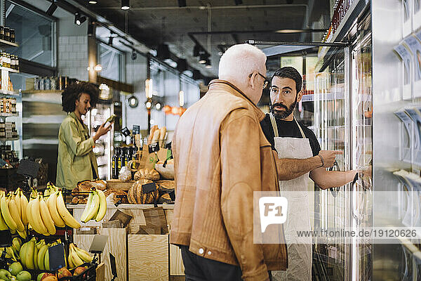 Älterer Einzelhandelsangestellter  der im Gespräch mit einem älteren männlichen Kunden im Lebensmittelgeschäft auf etwas zeigt