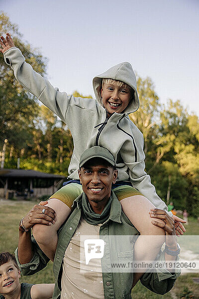 Portrait of male counselor carrying cheerful boy on shoulders while enjoying in playground