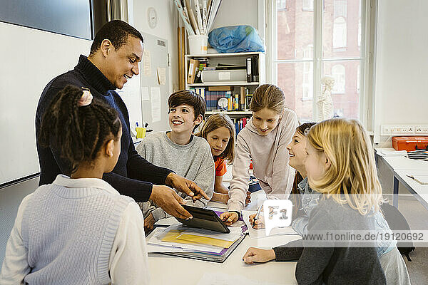 Männlicher Lehrer unterrichtet Schüler über einen Tablet-PC am Schreibtisch im Klassenzimmer