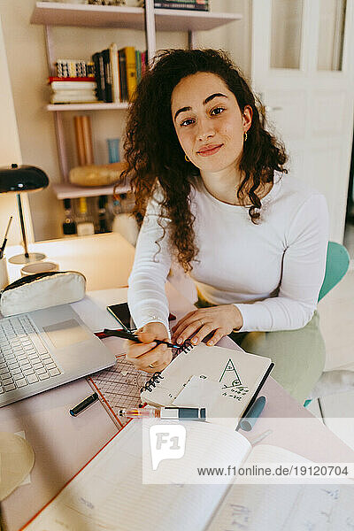 Porträt einer jungen Frau mit lockigem Haar  die am Schreibtisch Hausaufgaben macht