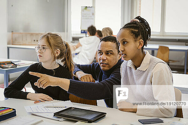 Männlicher Lehrer  der auf weibliche Schüler zeigt  die am Schreibtisch im Klassenzimmer sitzen
