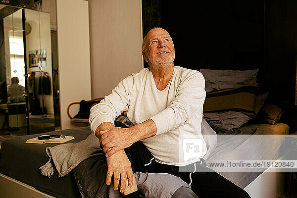 Lächelnder älterer Mann schaut auf  während er zu Hause auf dem Bett sitzt