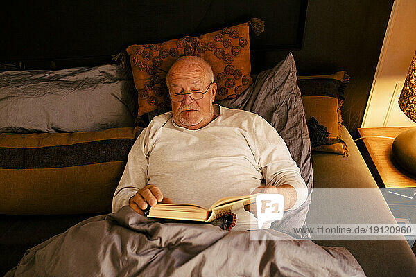 Älterer Mann liest ein Buch  während er zu Hause auf dem Bett liegt