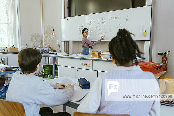 Smiling female teacher writing on whiteboard while teaching male students sitting in classroom