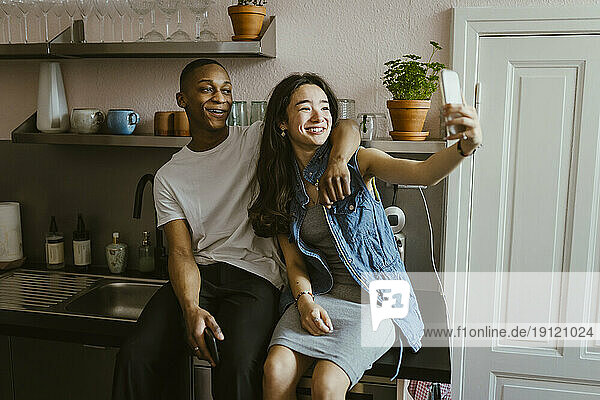 Glückliche junge Frau  die ein Selfie mit einem Freund macht  der auf der Küchentheke zu Hause sitzt