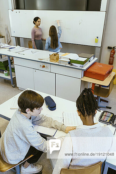 Hoher Blickwinkel eines Jungen  der einem männlichen Freund hilft  während er auf dem Schreibtisch im Klassenzimmer sitzt