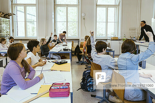 Männliche und weibliche Schüler heben die Hände  während sie auf Stühlen im Klassenzimmer sitzen