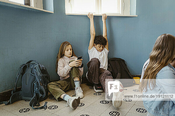 Mädchen  das ein Smartphone benutzt  neben einem männlichen Freund sitzend  der sich streckt  während er in der Schule sitzt