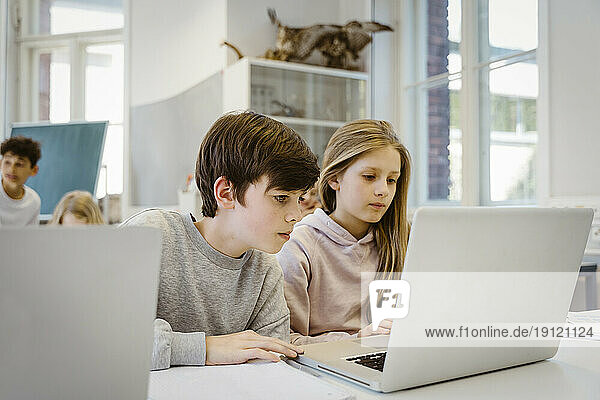 Konzentrierter Junge beobachtet Laptop mit Freundin am Schreibtisch im Klassenzimmer