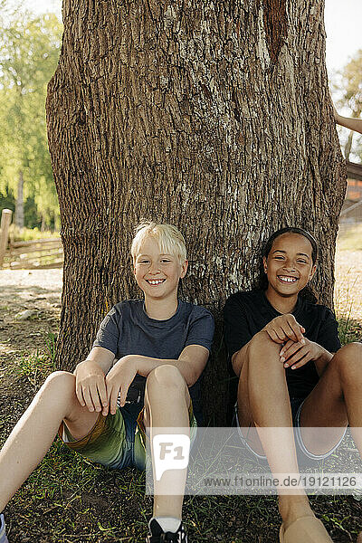 Portrait of smiling male and female friend sitting near tree trunk at summer camp