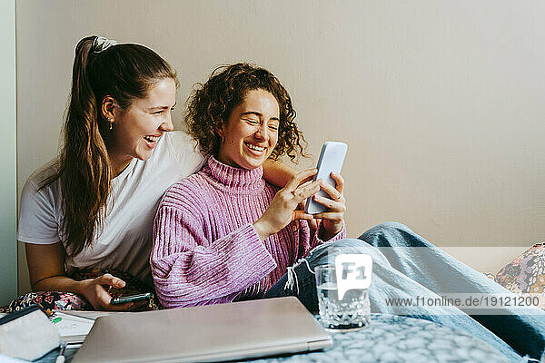 Fröhliche junge Freundinnen teilen sich ein Smartphone  während sie zu Hause sitzen