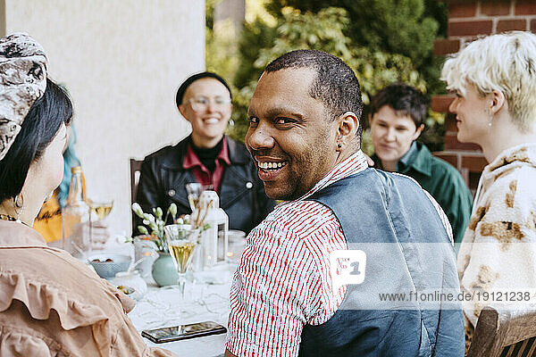 Portrait of smiling gay man looking over shoulder by friends during dinner party in back yard