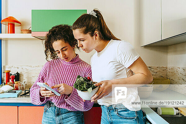 Junge Frau  die ihr Handy mit einer Freundin teilt  während sie sich zu Hause auf die Küchentheke stützt